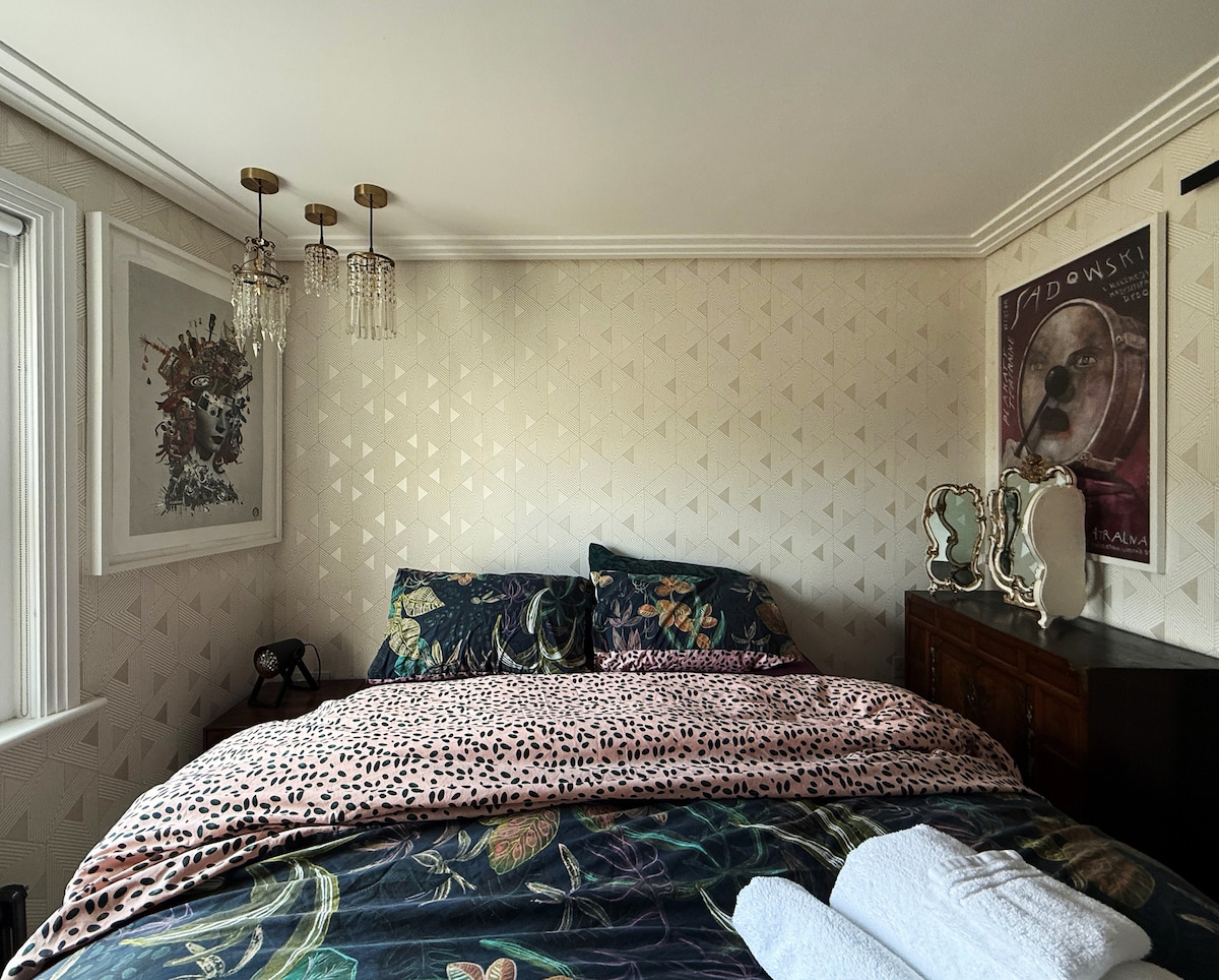 A cozy bedroom features a spacious bed adorned with a floral duvet and leopard print throw. Decorative artwork is displayed on the walls, accompanied by elegant light fixtures. A wooden dresser and a vintage mirror add a touch of charm, while natural light filters through the window.