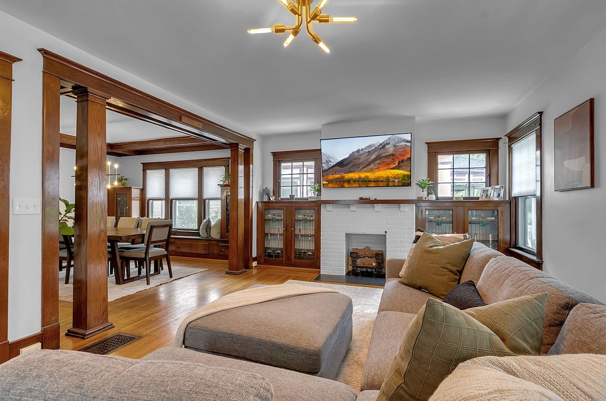 A spacious living room features natural woodwork and large windows allowing ample light. A comfortable sectional sofa, adorned with decorative pillows, faces a mounted television. A fireplace adds a cozy touch, while a dining area is visible in the background.