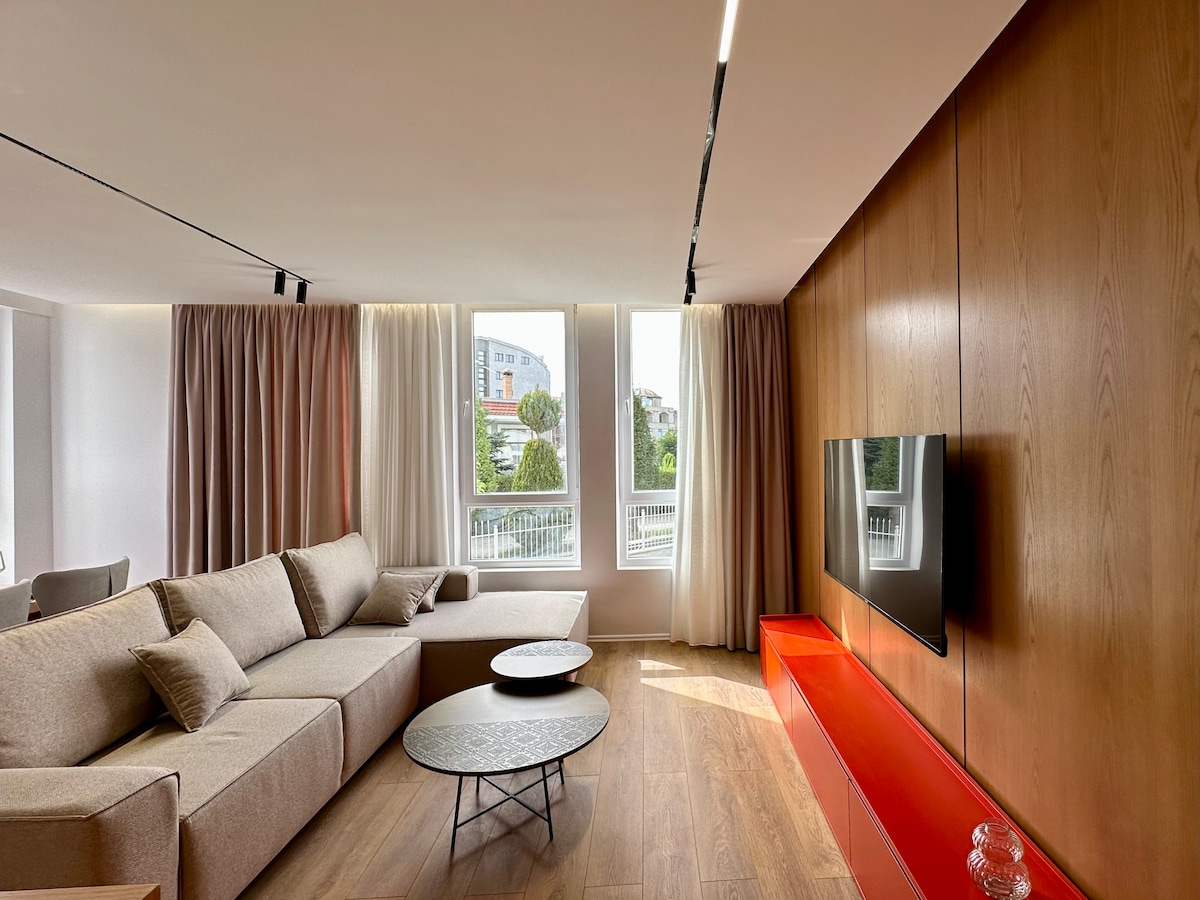 A modern living area features a spacious sectional sofa and two circular coffee tables. Large windows dressed with light curtains allow natural light to fill the room. A sleek red media console holds a flat-screen television against a wood-paneled wall, creating a contemporary atmosphere.
