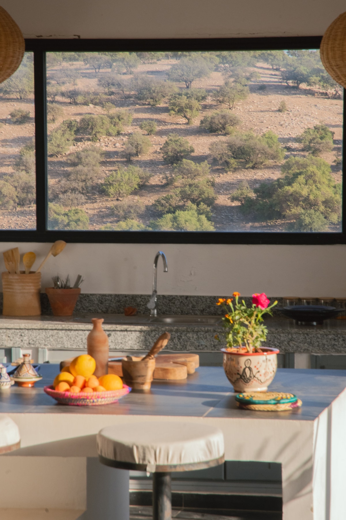 A modern kitchen area features a granite countertop with a decorative bowl and a pot. Fresh flowers are displayed in a ceramic vase, alongside a bowl of oranges. Large windows offer views of the surrounding hillside adorned with lush greenery.