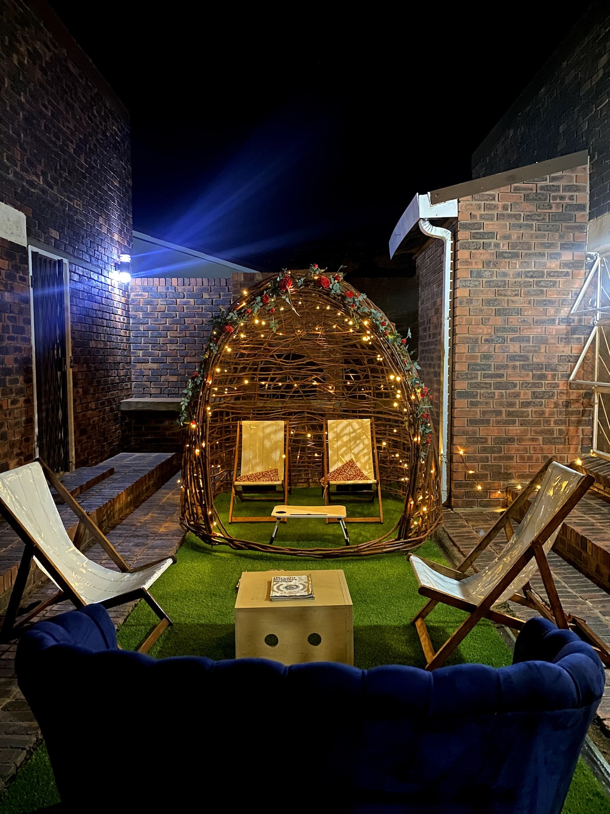 A cozy outdoor seating area is illuminated by strings of lights, featuring a decorative dome structure surrounded by four wooden chairs. Greenery covers the ground, and the warm glow adds a welcoming touch to the space.