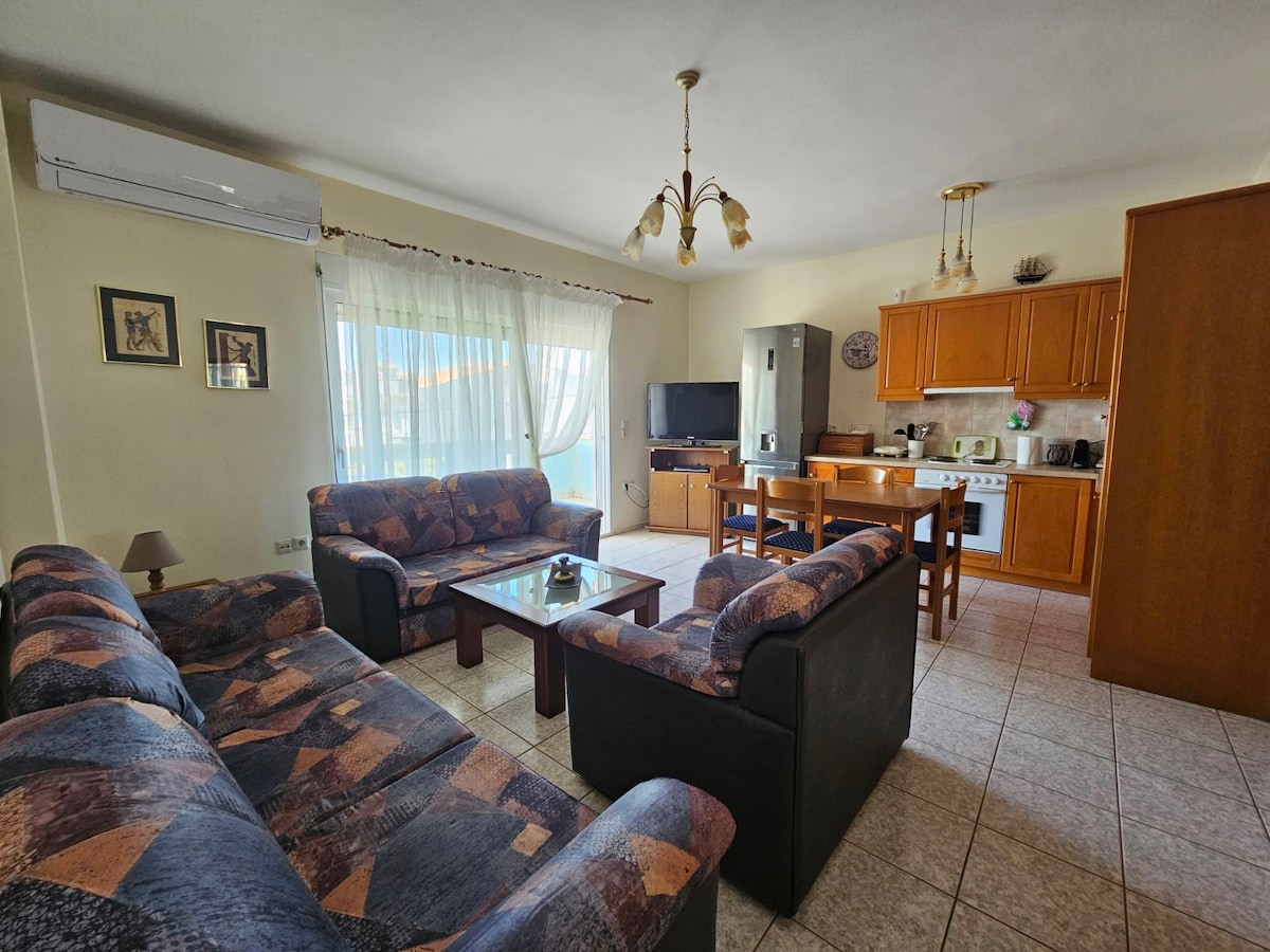 An inviting living area features comfortable seating arranged around a coffee table, with ample natural light coming through large windows. A dining table sits adjacent to a modern kitchen equipped with essential appliances, creating a functional and welcoming environment.