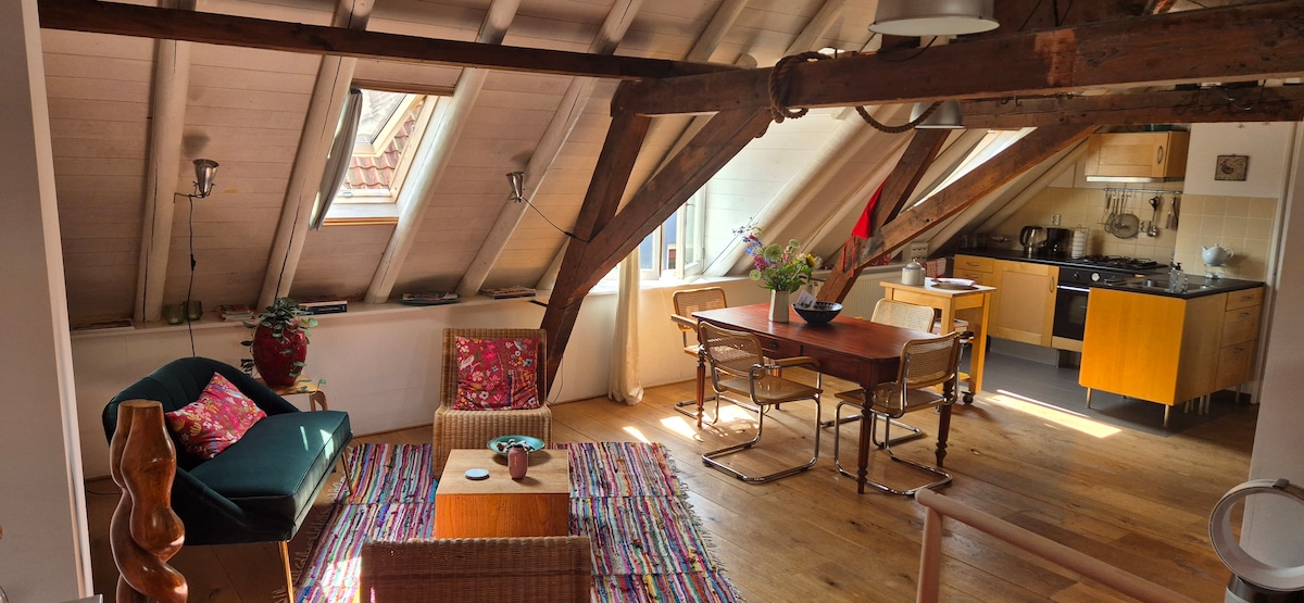 An airy loft space features exposed wooden beams and sloped ceilings. A cozy sitting area includes a green sofa and decorative pillows, while a dining table sits across from a fully equipped kitchen. Natural light streams in from the windows, enhancing the warm wooden floors.