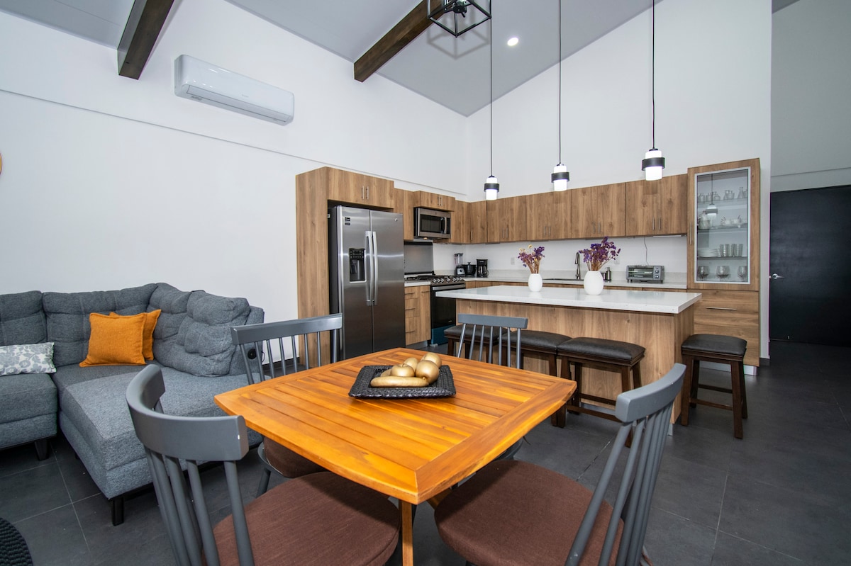 A modern kitchen and dining area are featured, showcasing a wooden table with a bowl of decorative items. Comfortable seating is arranged around the table, while the kitchen includes stainless steel appliances and a sleek design. Natural light fills the space through large windows.