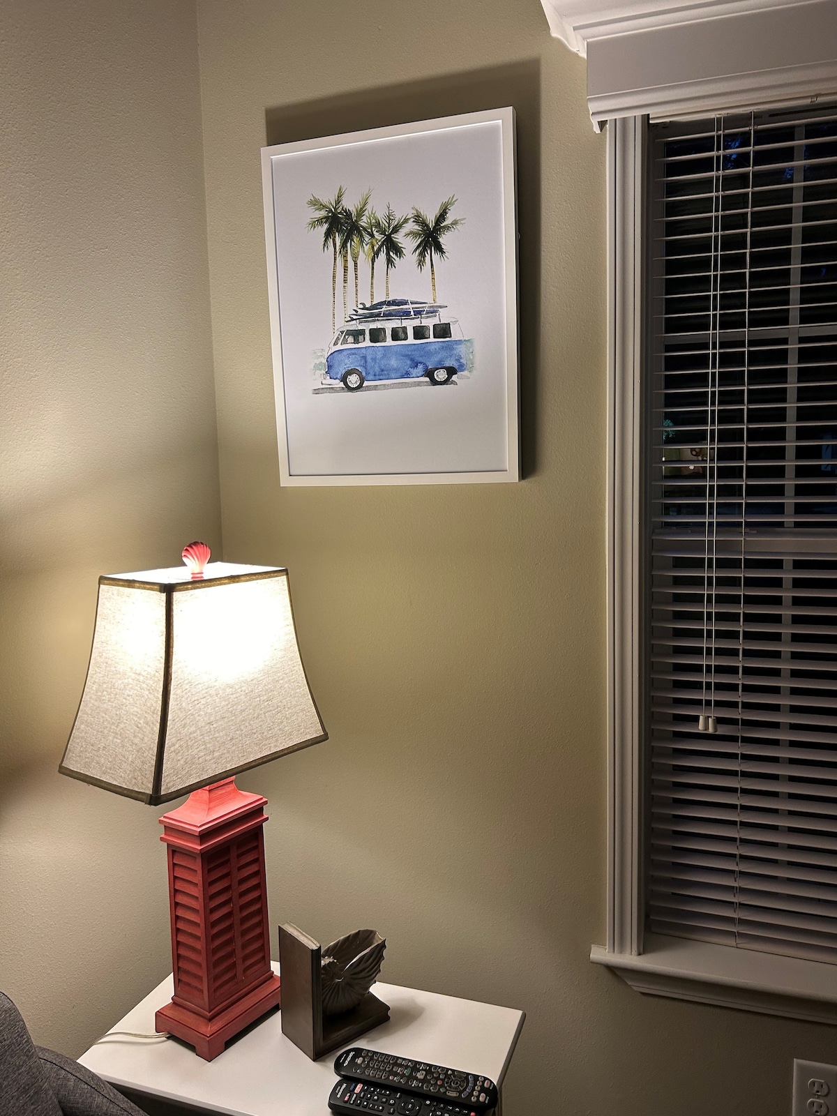 A corner of the living room is highlighted by a framed artwork featuring a blue vintage van surrounded by palm trees. A small lamp with a light shade sits on a white side table, casting a soft glow. Blinds partially cover a nearby window.