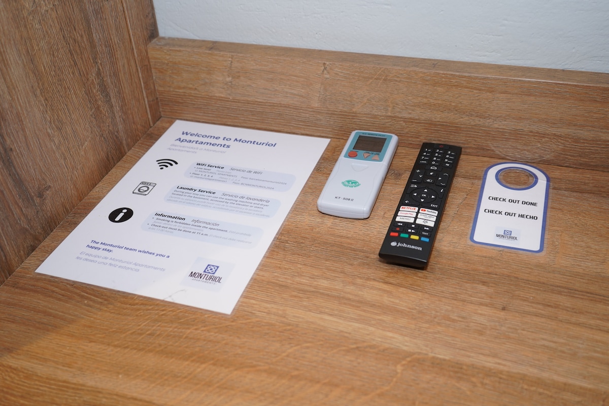 A wooden countertop displays a welcome document outlining services and amenities, alongside a remote control for the TV. Additional items include a light control panel and a door hanger for check-out instructions, all arranged neatly for guest convenience.