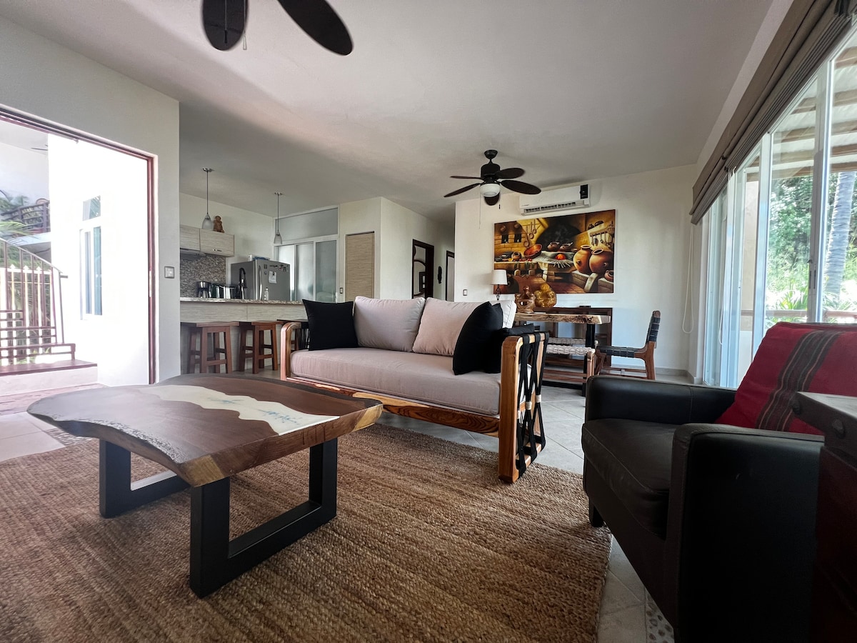 A cozy living room features a mix of seating options, including a light-colored sofa and a dark armchair, arranged around a stylish coffee table. Natural light streams in through large sliding glass doors, enhancing the open layout that connects to the dining area and kitchen.