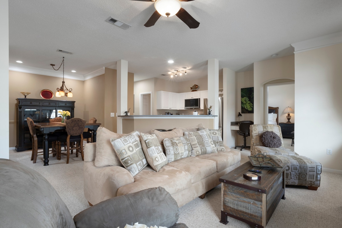A spacious living area features a large sectional sofa adorned with decorative pillows, complemented by a wooden coffee table. In the background, a dining area is visible with a dark wooden table and chairs. The kitchen offers a clear view with modern appliances against light-colored cabinetry.