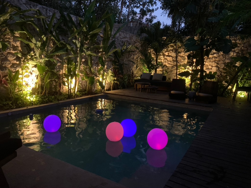 A private pool is surrounded by lush tropical plants and a stone wall, illuminated by soft lighting. Colorful floating orbs in shades of purple, pink, and white add a gentle glow to the water. Comfortable lounge chairs are positioned nearby for relaxation.