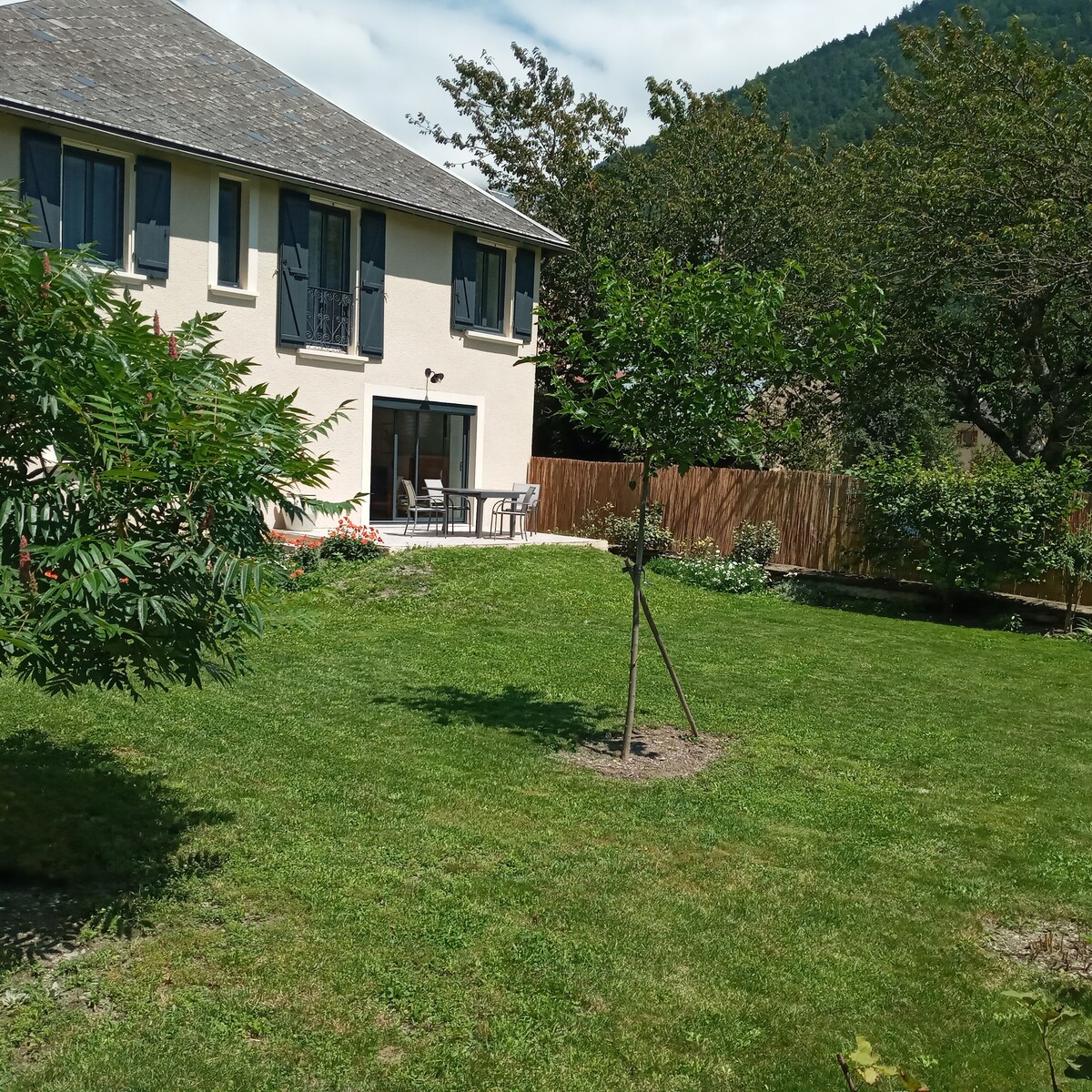 The exterior of the house is surrounded by a well-maintained garden featuring lush green grass and young trees. A patio area with outdoor seating is visible, providing a cozy space for relaxation. The backdrop consists of gently sloping hills and a partly cloudy sky.