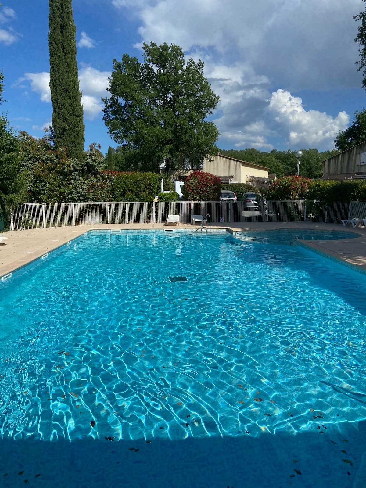 A clear blue swimming pool is surrounded by a fenced area with lounge chairs positioned along the perimeter. Lush greenery, including trees and shrubs, frames the pool, creating a serene and inviting environment. The sky is partially cloudy, enhancing the peaceful atmosphere.