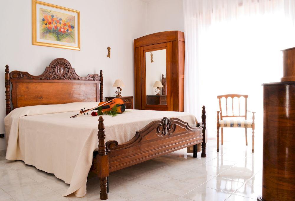 A spacious bedroom features a wooden bed frame with intricate detailing and a neutral bedspread. A violin rests on the bed. A large mirror and wooden wardrobe reflect natural light from a nearby window, complemented by a chair with a classic design.