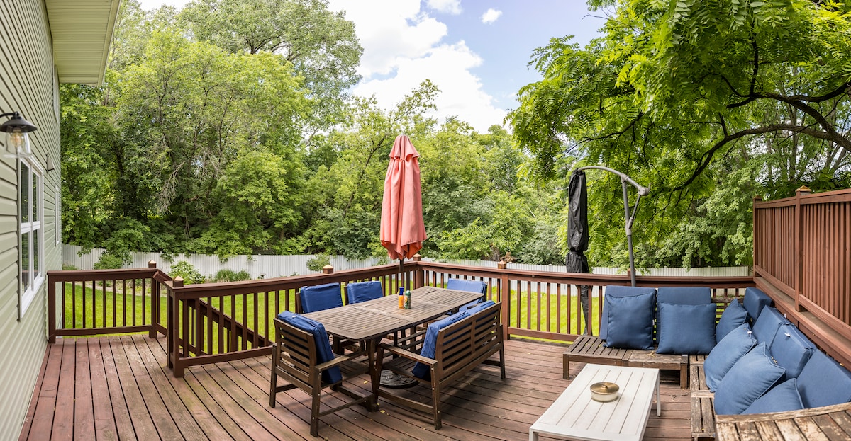 An outdoor deck is displayed with wooden flooring and a spacious arrangement of seating. A large dining table is accompanied by cushioned chairs. An umbrella offers shade, while green trees create a natural backdrop, enhancing the serene ambience of the outdoor space.
