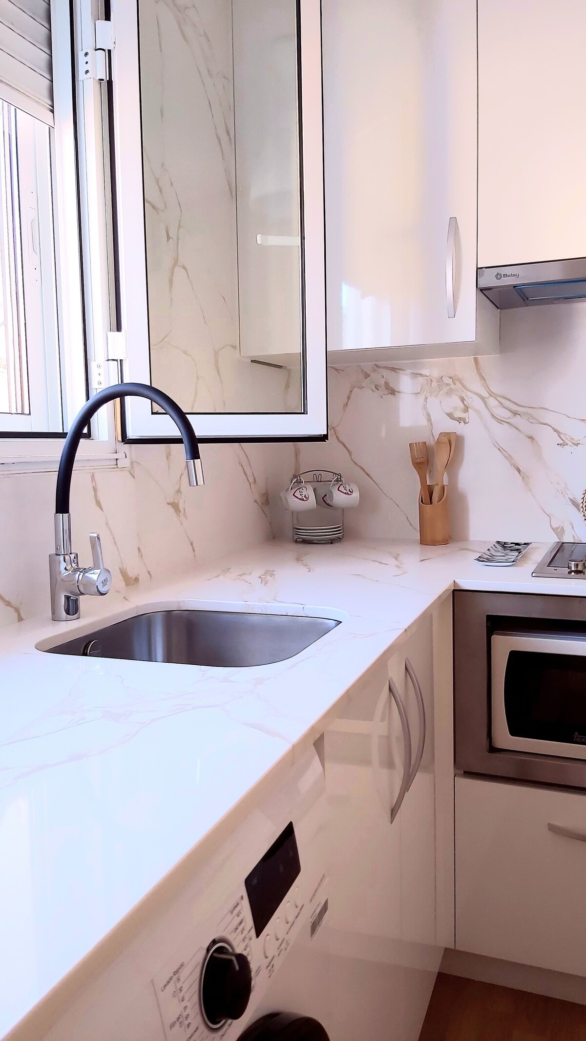 A modern kitchen area is highlighted by sleek, white cabinetry and a polished, marble countertop. A stainless steel sink is positioned beneath a window, allowing natural light to illuminate the space. Essential kitchen appliances, including a stovetop and microwave, are neatly integrated.