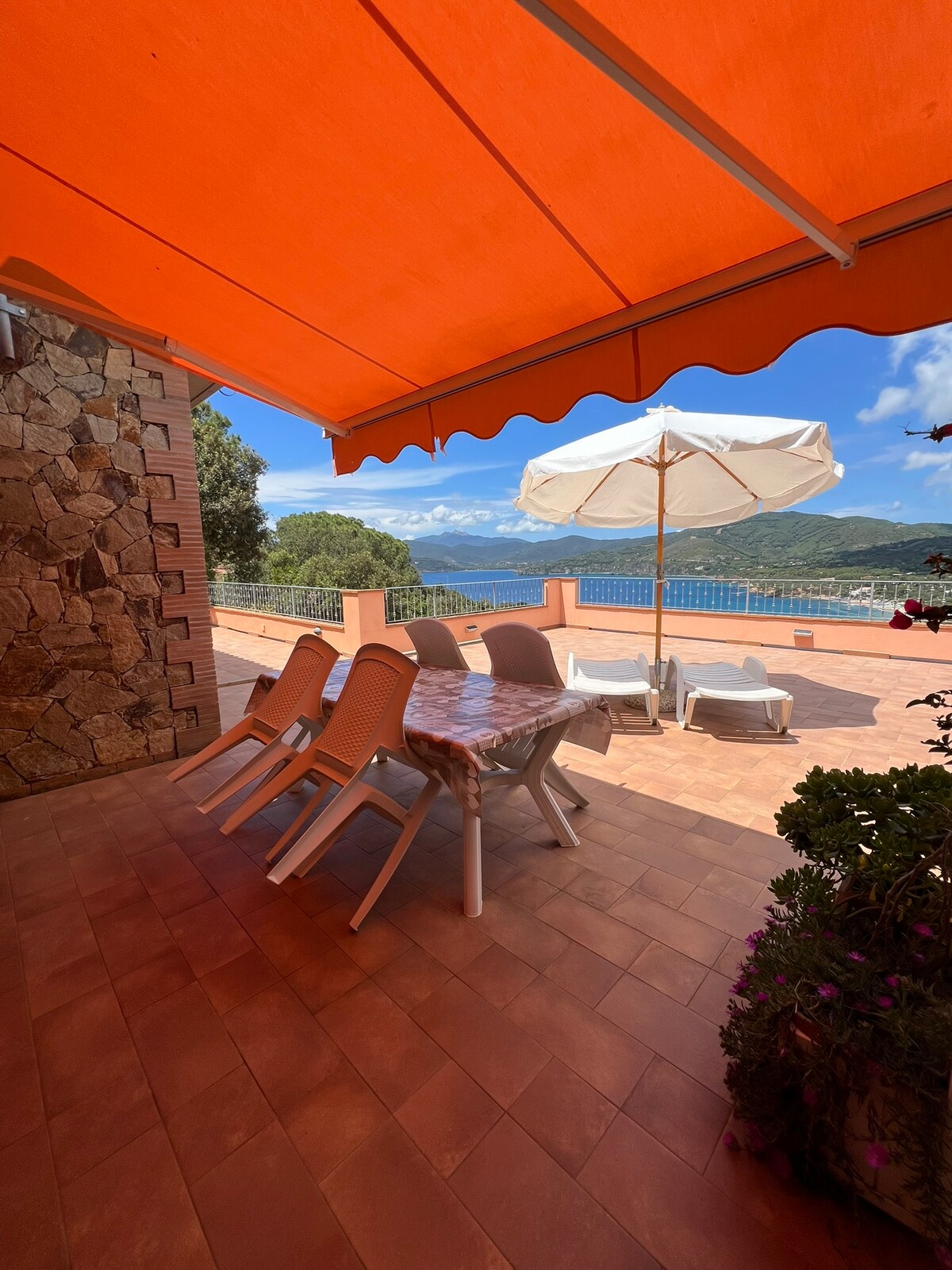 An outdoor terrace features a large orange awning providing shade over a dining area. A round table is set with chairs, while sun loungers and a large white umbrella are positioned nearby, offering views of the landscape and water below.