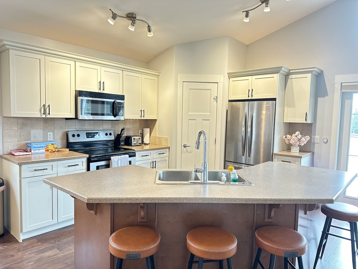 A modern kitchen showcases light-colored cabinetry and stainless steel appliances. A large island with seating for four is complemented by a double sink. Natural light streams in from a nearby window, highlighting the spacious layout and counter space.