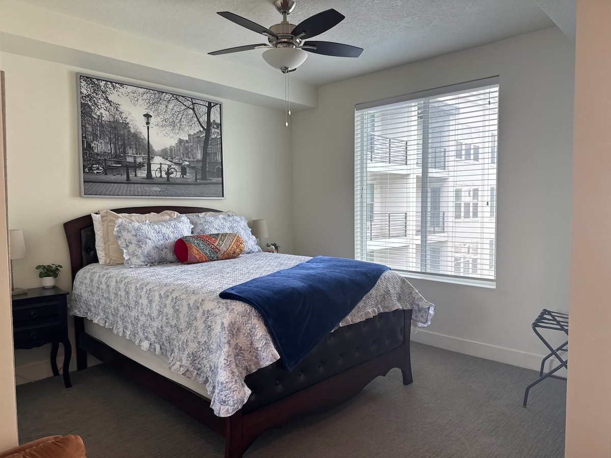 A comfortable bedroom features a spacious bed adorned with patterned bedding and plush cushions. A contemporary ceiling fan is mounted above, while natural light filters through the window adorned with blinds. A small nightstand stands beside the bed, enhancing the minimalist design.