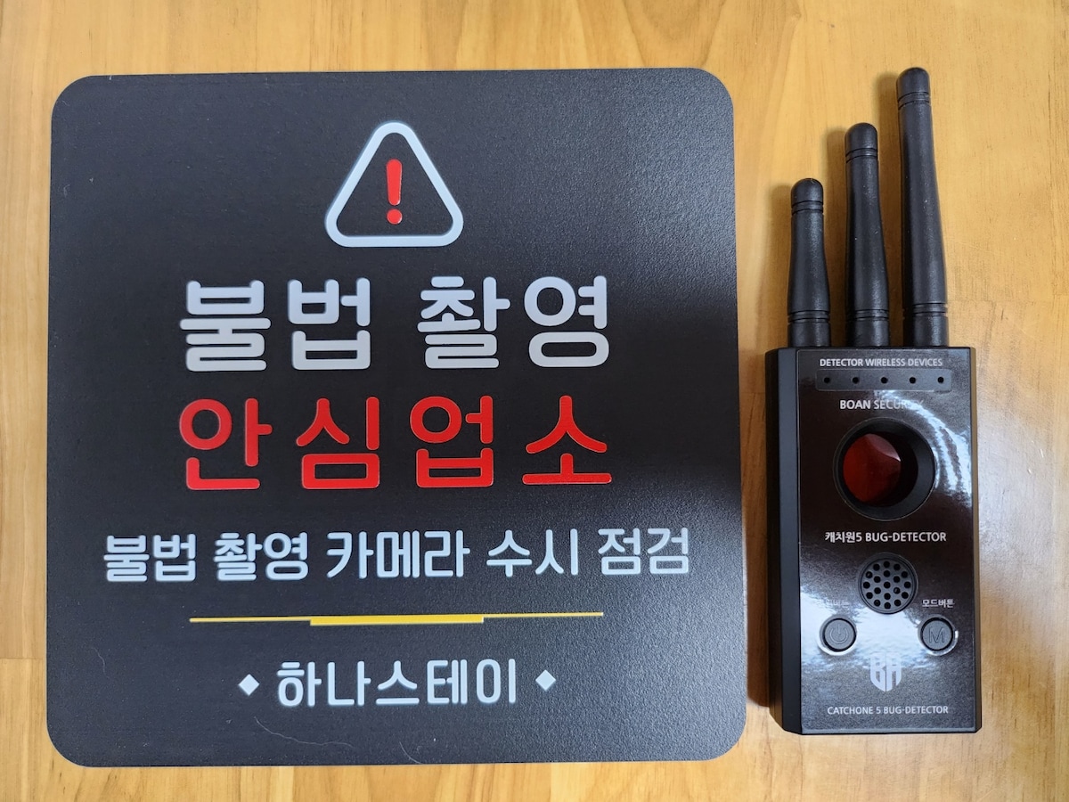 A bug detector device is displayed on a wooden surface, accompanied by a warning sign in Korean text. The device features three antennas and a central button, while the sign highlights the establishment’s focus on privacy and security.