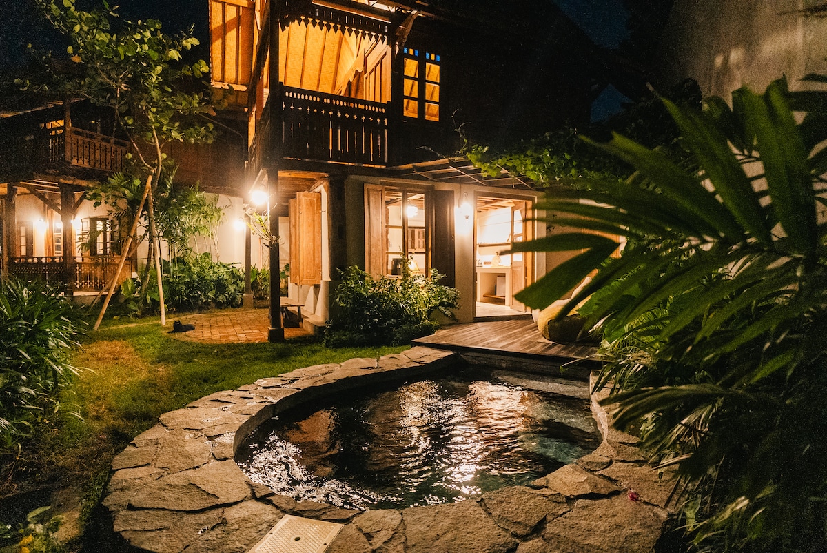 A private plunge pool is nestled among lush greenery, surrounded by stone paving and illuminated by soft outdoor lighting. The two-story luxury cottage features wooden balconies and large windows, offering views of the garden and inviting an open-air ambiance.