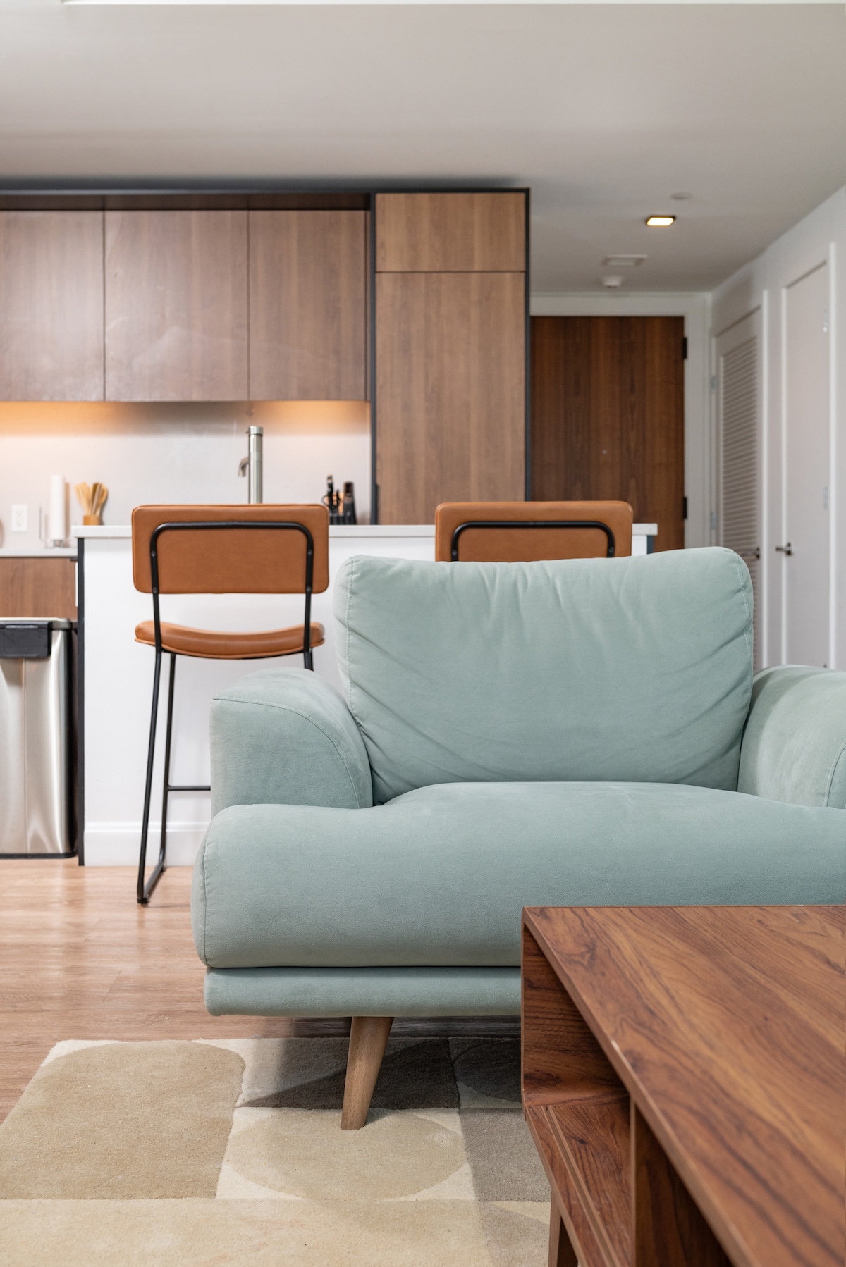 A cozy living area is showcased, featuring a soft, pastel green armchair with wooden legs. A wooden coffee table sits in front, complemented by the sleek kitchen area in the background, highlighted by modern cabinetry and bar stools.