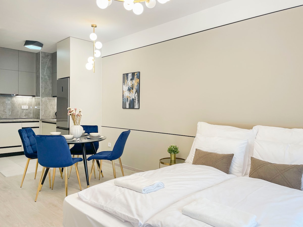 A modern living area is presented with a spacious double bed and a dining table for four. Soft white bedding complements the neutral walls. A stylish light fixture hangs above, while a small decorative plant adds a touch of greenery. The kitchen area, partially visible, features contemporary cabinetry.