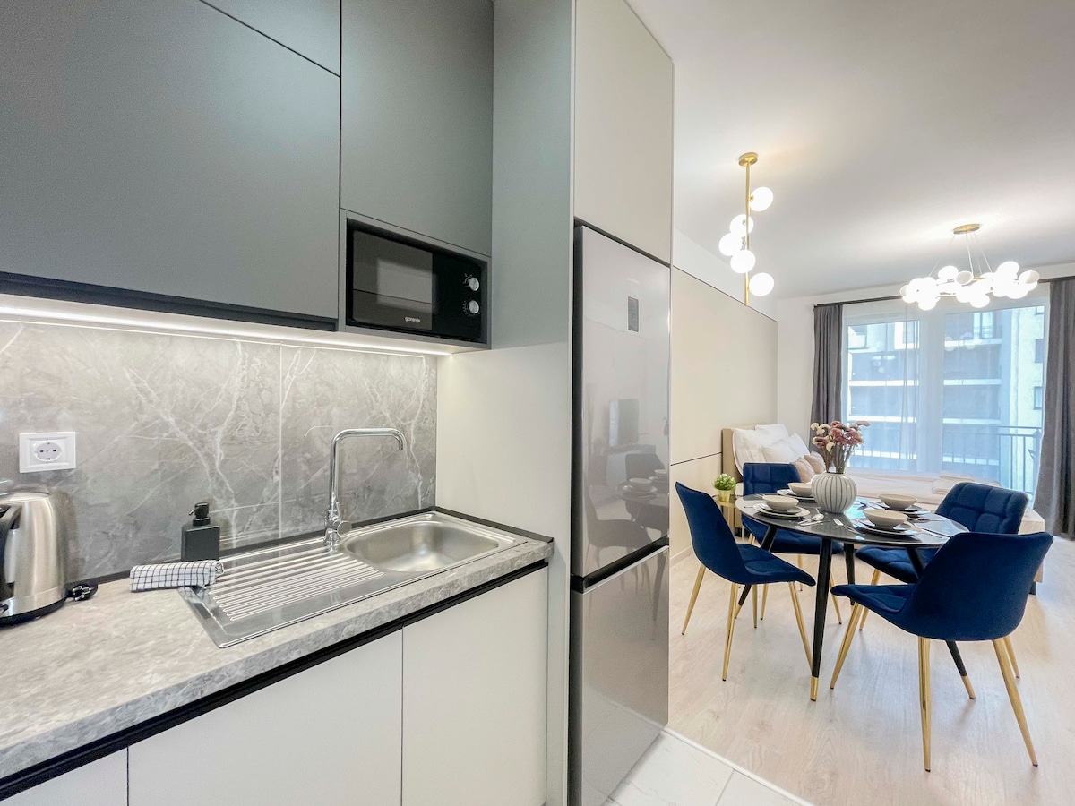 A modern kitchen area features sleek cabinetry and a gray countertop. Stainless steel appliances, including a kettle and microwave, are visible. Adjacent to the kitchen is a dining space with a round table surrounded by four blue chairs. Natural light floods the room through large windows.