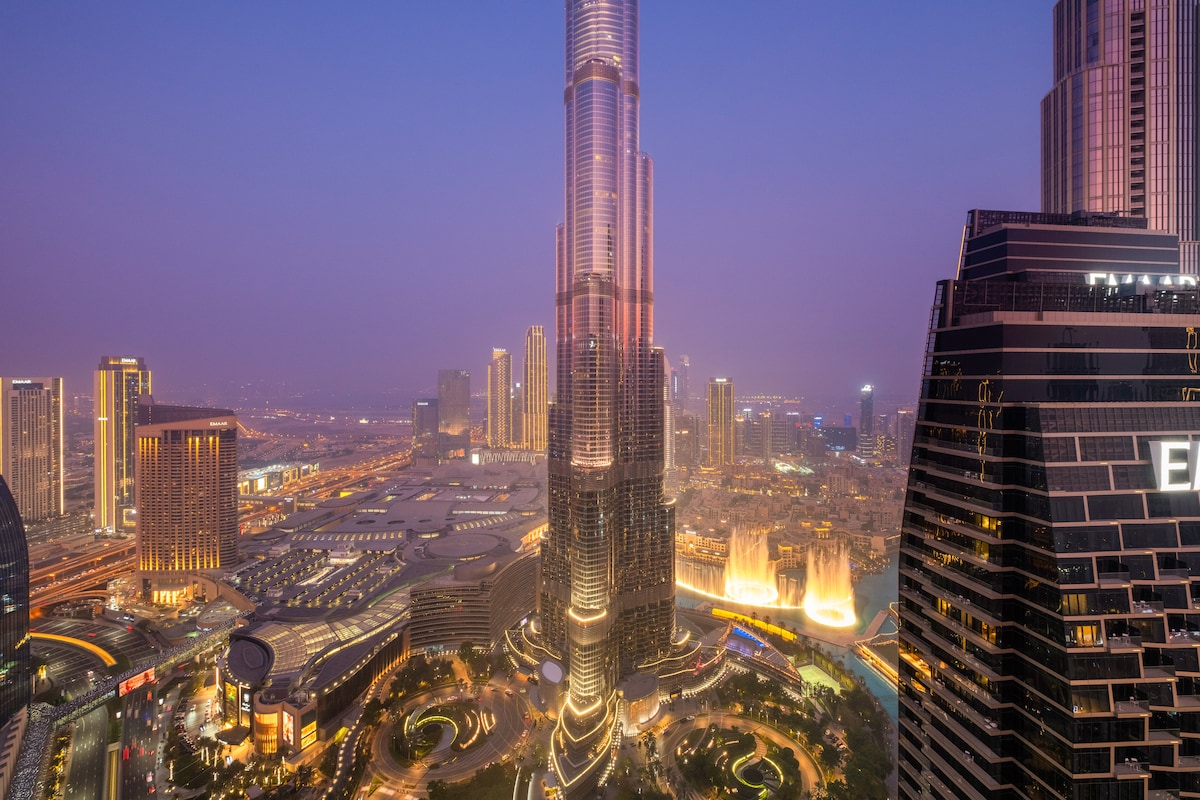 A high-angle view captures the Burj Khalifa towering over the cityscape at dusk. The surrounding buildings and vibrant lights reflect the energy of Dubai, while the illuminated fountain at the base creates a focal point, blending seamlessly into the urban landscape.