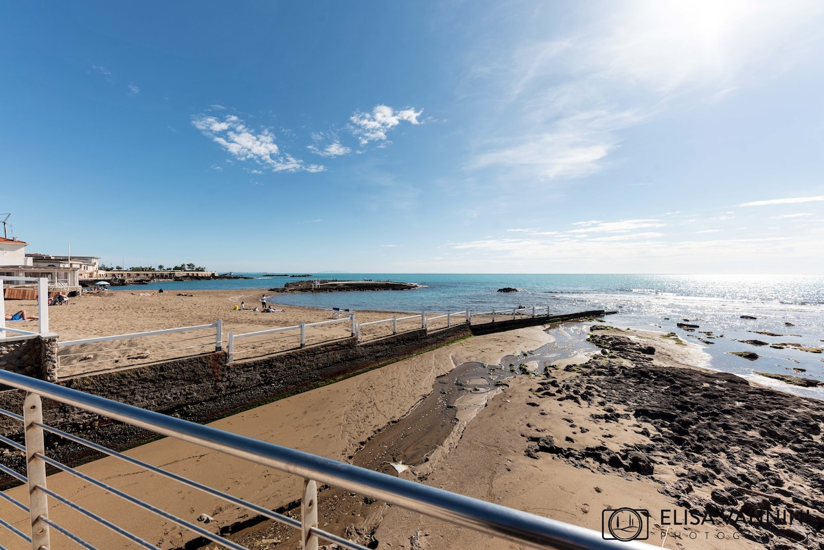 A shoreline view captures a sandy beach leading to a calm sea. The expanse of water reflects the clear blue sky with soft clouds, while a wooden path extends into the water. Rocky formations are visible along the coastline, enhancing the natural scenery.