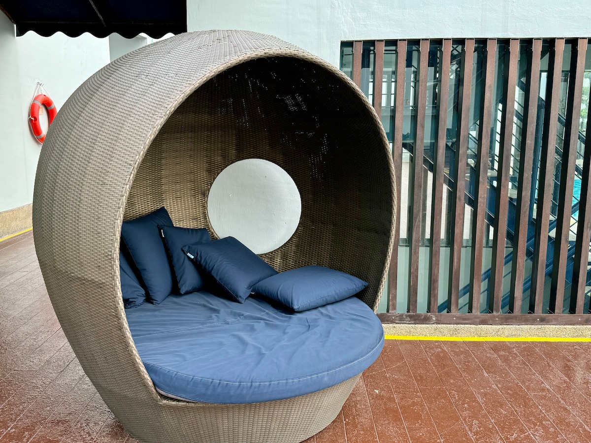 A round, woven seating area is positioned outdoors, featuring a soft blue cushion and several navy throw pillows. The structure offers a cozy space for relaxation, framed by walls with horizontal slats in a contrasting dark wood finish.