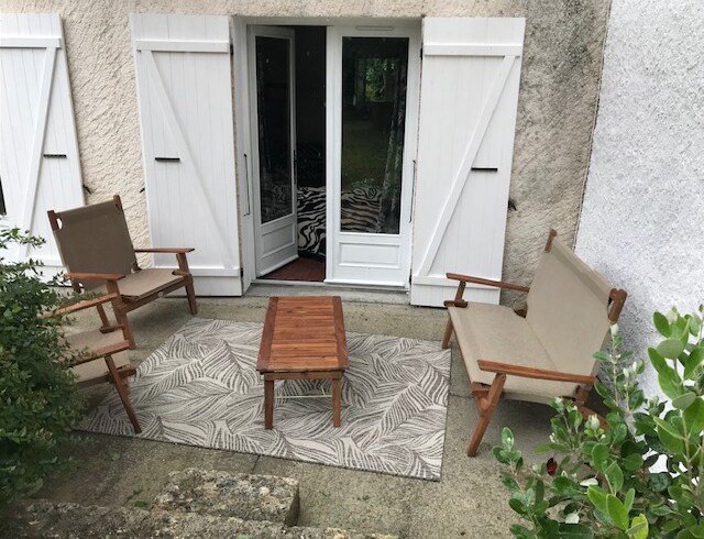 An outdoor seating area is presented, featuring two armchairs and a small coffee table, all set on a patterned rug. Double doors open to an interior space, with greenery bordering the area, providing a serene and inviting ambiance.