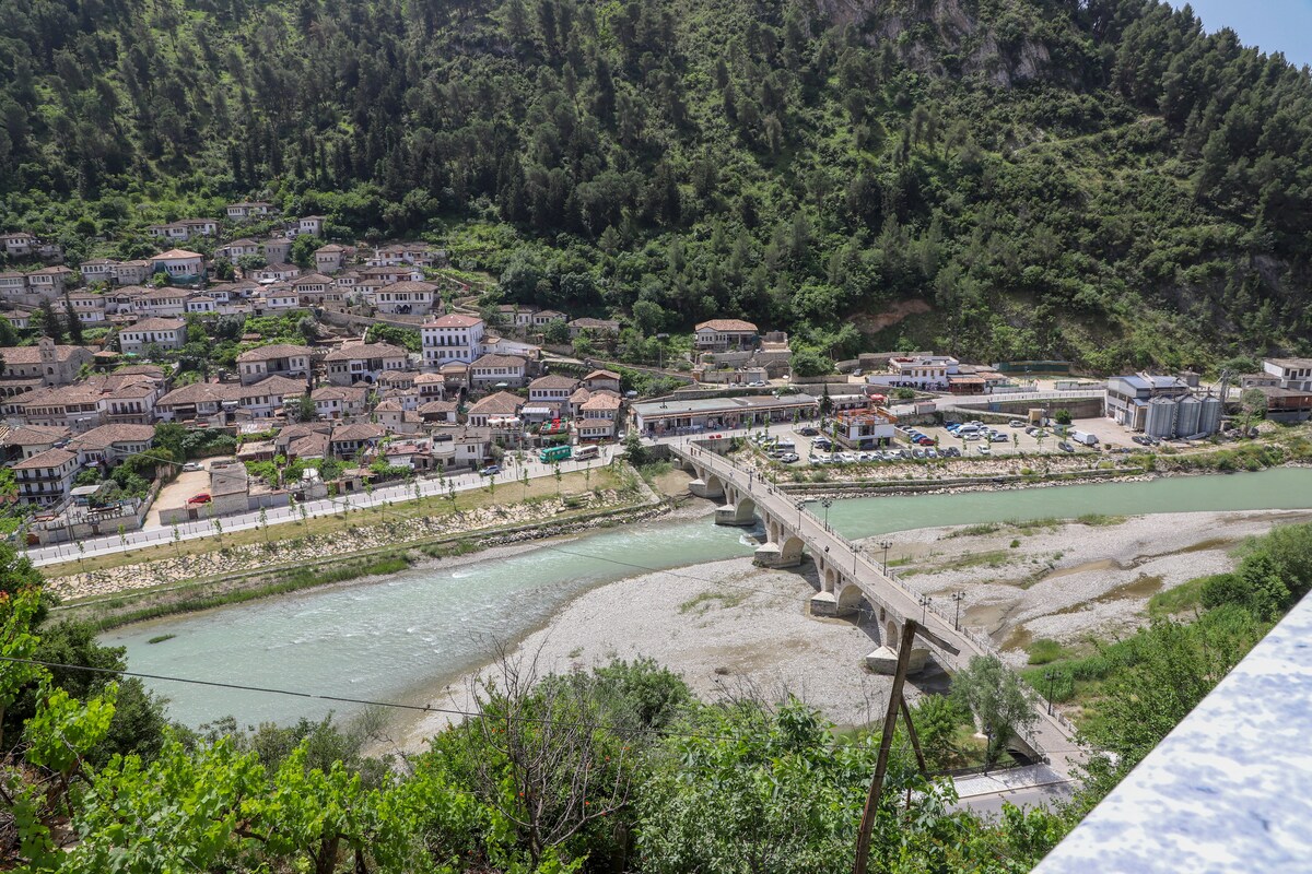 A panoramic view of the Gorica quarter shows a cluster of traditional houses with tiled roofs nestled along the lush hillside. The Gorica Bridge spans the light green Osumi River, with the valley and surrounding mountains providing a natural backdrop.