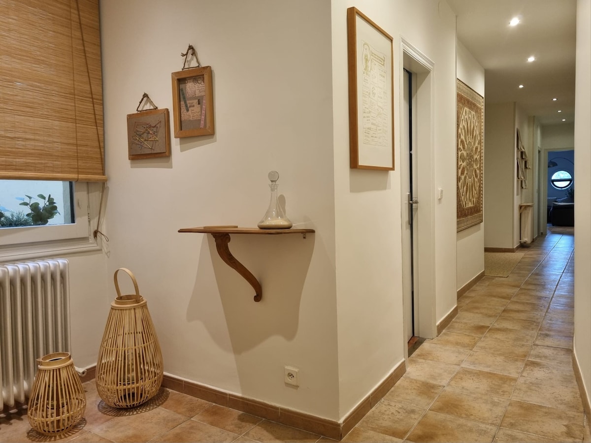 A hallway is presented with earthy tiles and neutral-colored walls. A wooden shelf is mounted on the wall, displaying a decorative glass item. Light spills in from a window adorned with bamboo blinds, and a textured wall hanging adds visual interest to the space.