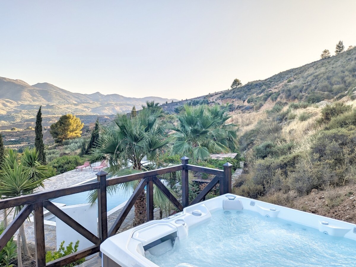 A hot tub is situated on a wooden deck, surrounded by lush greenery and palm trees. The background features rolling hills and distant mountain ranges under a clear sky, offering a serene and private outdoor setting.