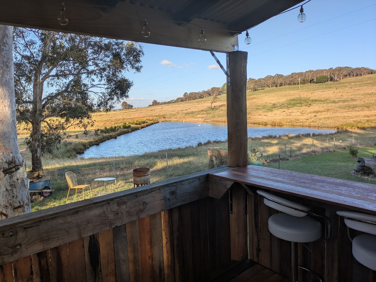 A view from the tiny house reveals a serene dam surrounded by grassy fields. The image captures a rustic wooden bar with a panoramic window, offering a glimpse of the landscape dotted with native trees and a few chairs positioned nearby.