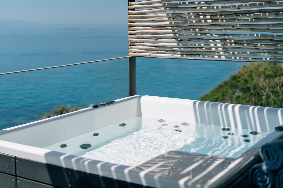A private jacuzzi is positioned on a spacious terrace, surrounded by vertical wooden slats that provide partial privacy. The tranquil water surface reflects the sunlight, while the expansive view of the sea stretches out in the background.