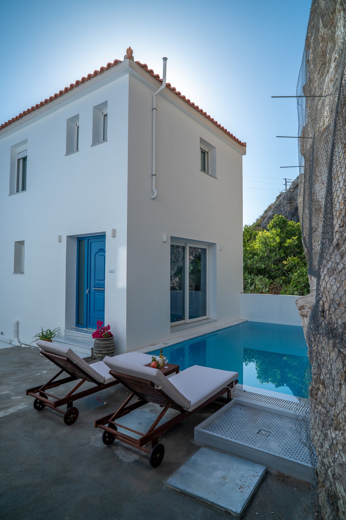 A modern villa features a bright blue door and large windows that open to an inviting pool area. Two wooden loungers with cushions are arranged beside the shimmering pool, creating a tranquil outdoor retreat against a natural rock backdrop.