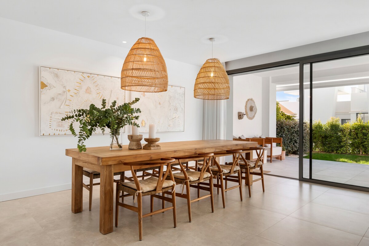 A wooden dining table is surrounded by six chairs, with natural light pouring in from large glass doors. Two woven pendant lights hang above the table, where a decorative arrangement of greenery and candles is displayed. A lush outdoor area is visible through the glass doors.