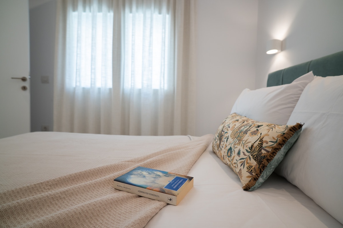 A neatly arranged sleeping area features a spacious bed layered with crisp white linens and a decorative pillow. A light blanket is draped over the bed, and a book rests on the covers. Natural light filters through sheer curtains, creating a serene atmosphere.