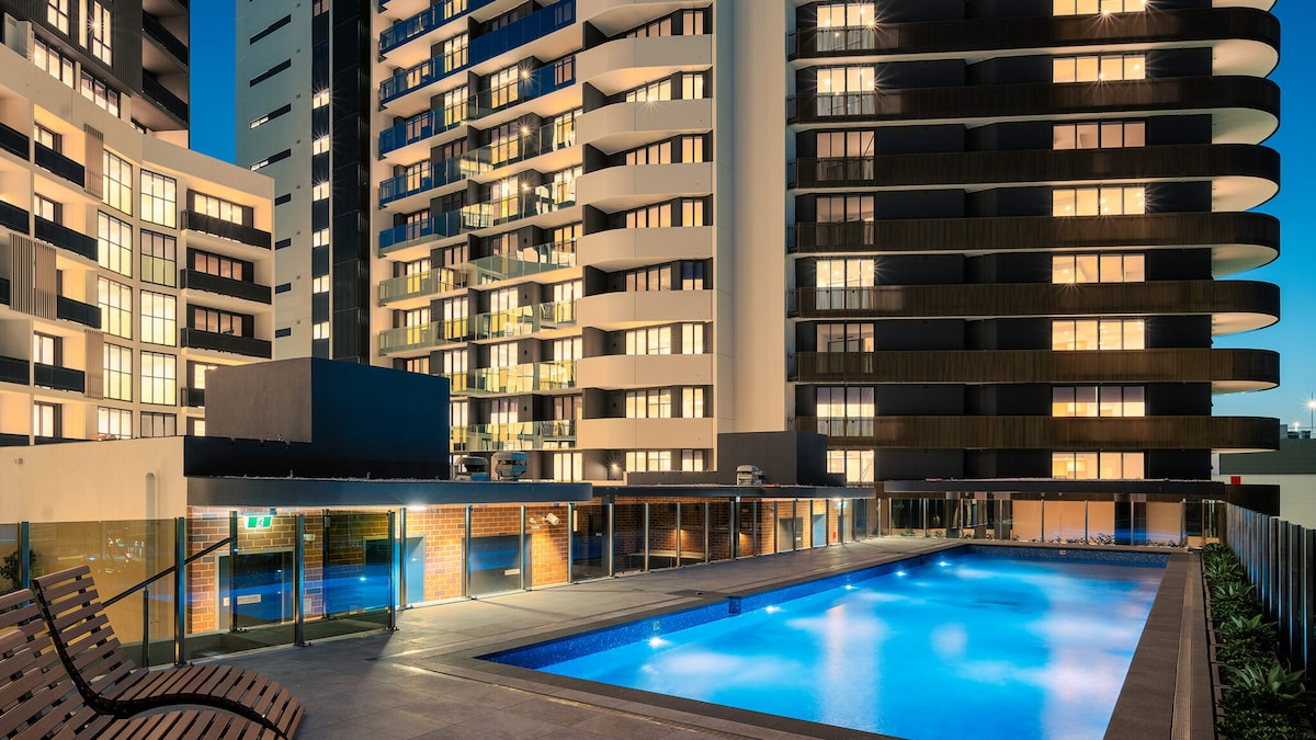 A modern apartment complex is illuminated at night, showcasing a sleek outdoor pool surrounded by a spacious deck. The building’s exterior features a combination of glass and dark materials, creating a contemporary urban atmosphere.