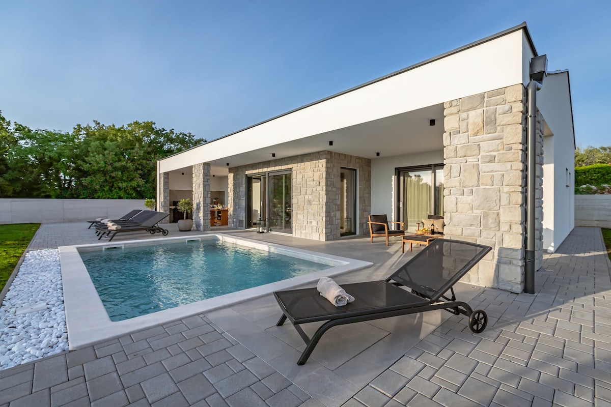 A modern villa exterior features a private swimming pool with deckchairs positioned nearby. The open terrace area includes a casual seating arrangement. The facade combines smooth white walls with natural stone, and large glass doors provide a view of the inviting outdoor space.