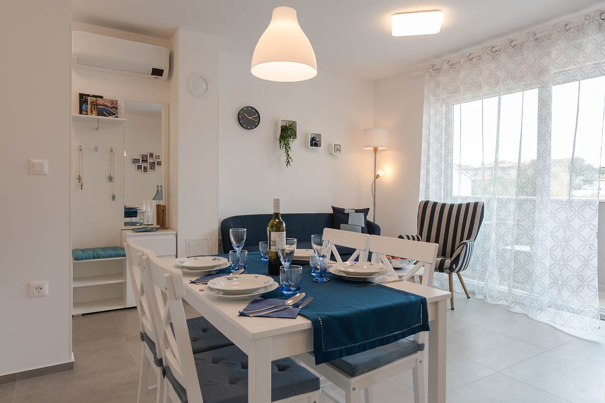 A bright dining area is showcased with a white table set for four, complemented by blue accents. A navy sofa provides additional seating nearby. Large windows with sheer curtains allow natural light to fill the space, while a contemporary light fixture hangs above the table.