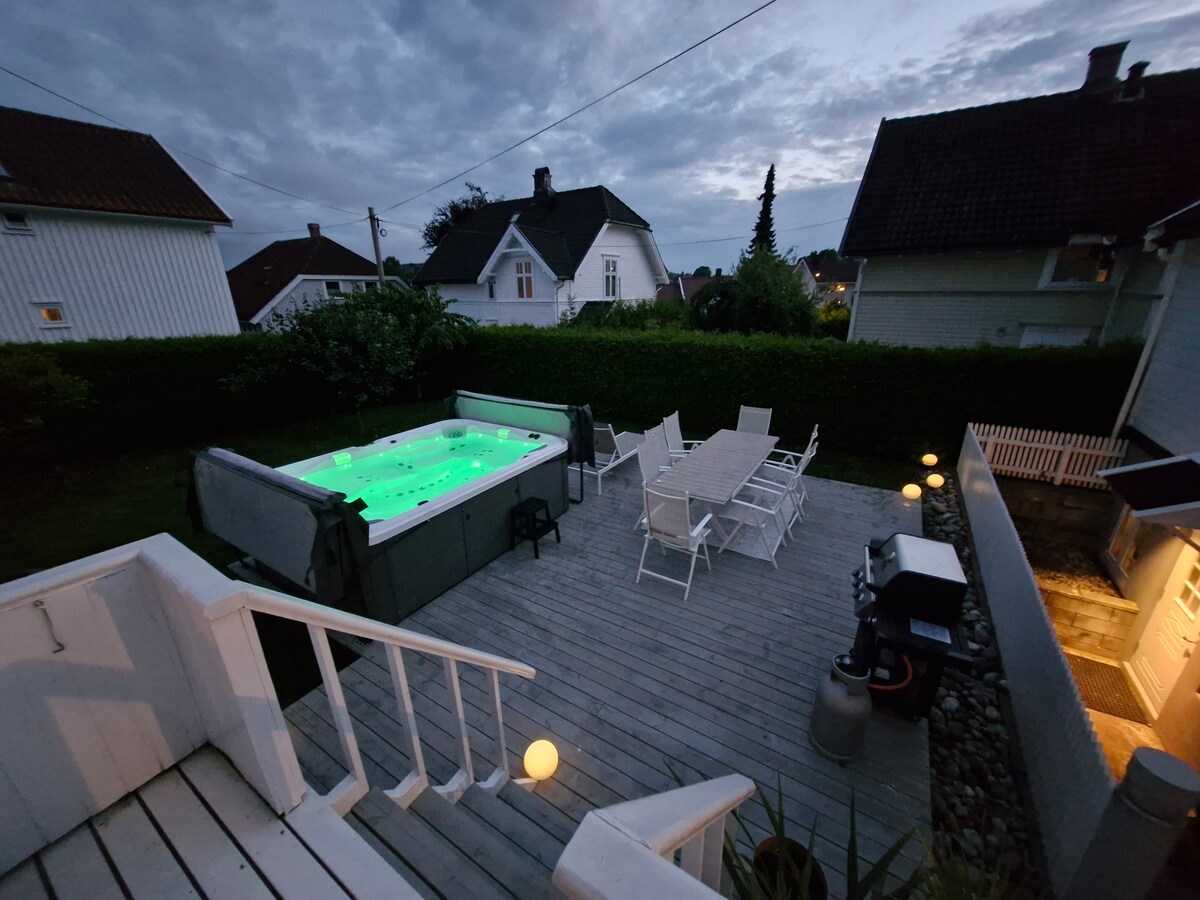 A well-maintained outdoor area features a jacuzzi illuminated in green, surrounded by wooden decking. A spacious dining table with several chairs is positioned nearby, with outdoor lighting and a barbecue grill visible. Lush greenery and neighboring houses complete the serene setting.