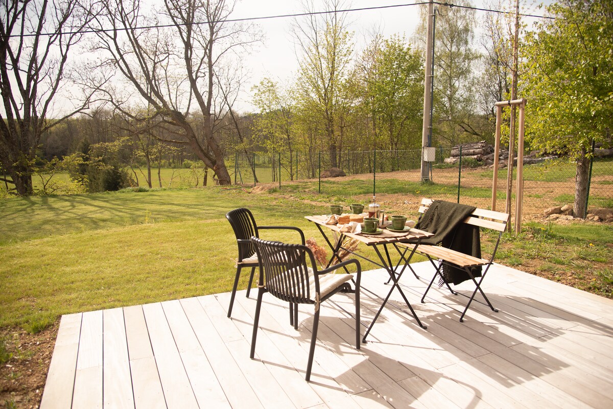 A patio area is visible, featuring two black chairs and a small folding table set for an outdoor meal. Lush green grass surrounds the space, and trees can be seen in the background, providing a serene natural setting.