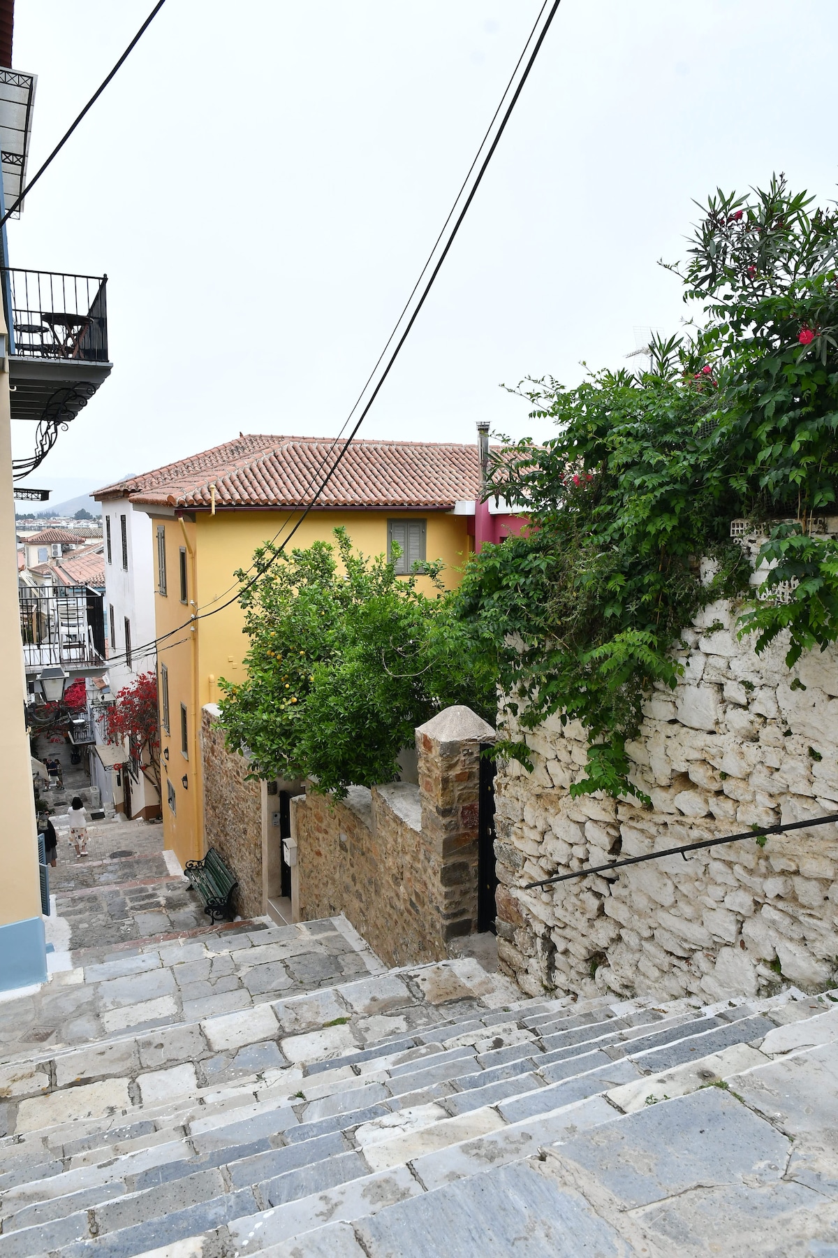 A quiet street in the old town features traditional stone steps leading downward. Colorful houses with tiled roofs are framed by lush greenery and blooming flowers, evoking a charming, historic atmosphere. The cobblestone path is bordered by rustic walls, reflecting the character of the surroundings.