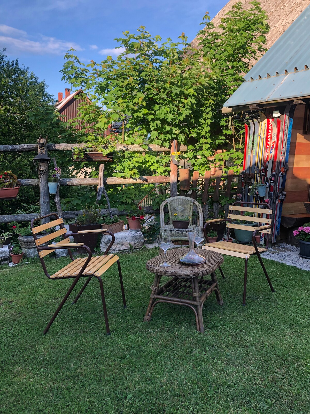 A small outdoor seating area is displayed, featuring two wooden chairs with metal frames and a woven table in the center. Lush greenery surrounds the space, complemented by vibrant plants in pots. A rustic fence adds to the charming environment.