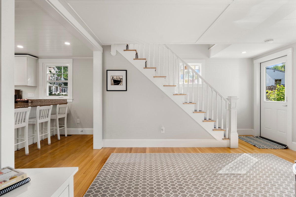 A bright and airy open-concept living area features light-colored walls and warm wood flooring. A staircase with white railings leads to the upper level, while a cozy kitchen bar with seating for four is visible in the background. Natural light streams in through the nearby door.