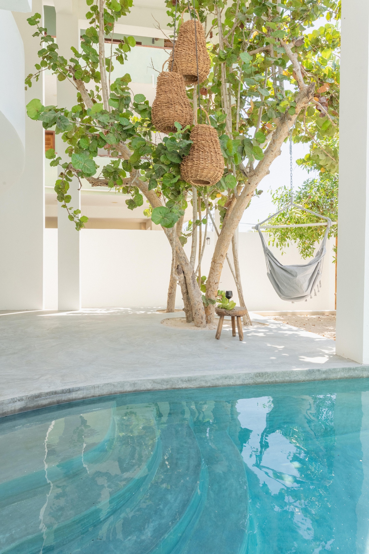 A tranquil space features a curved private pool reflecting the surrounding greenery. A majestic tree with hanging woven baskets and a comfortable hammock adds a serene touch, while a small wooden stool sits beside the water, enhancing the inviting atmosphere.