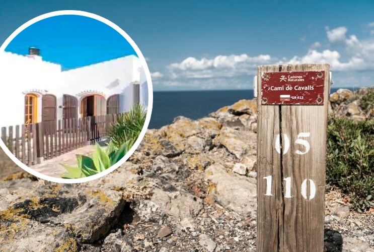 A wooden sign with white numbers indicates the location on the Cami de Cavalls trail, accompanied by a view of rocky terrain and the sea. The image features a charming house with white walls and arched windows in the background, framed by green plants.