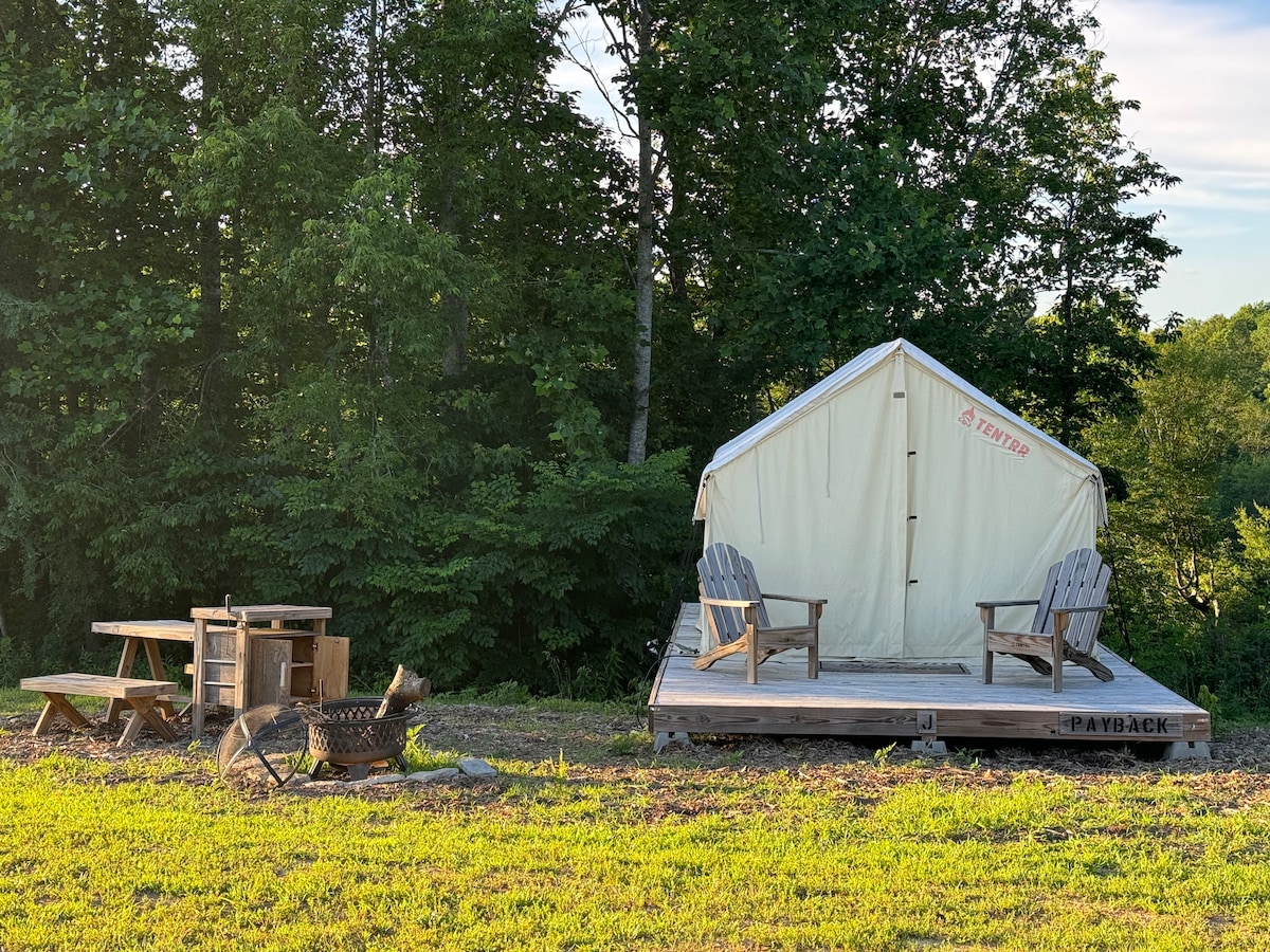 A canvas glamping tent sits on a raised wooden deck surrounded by greenery. Two Adirondack chairs are positioned in front, adjacent to a picnic table and a fire pit equipped with a grill grate, inviting a relaxing outdoor experience.