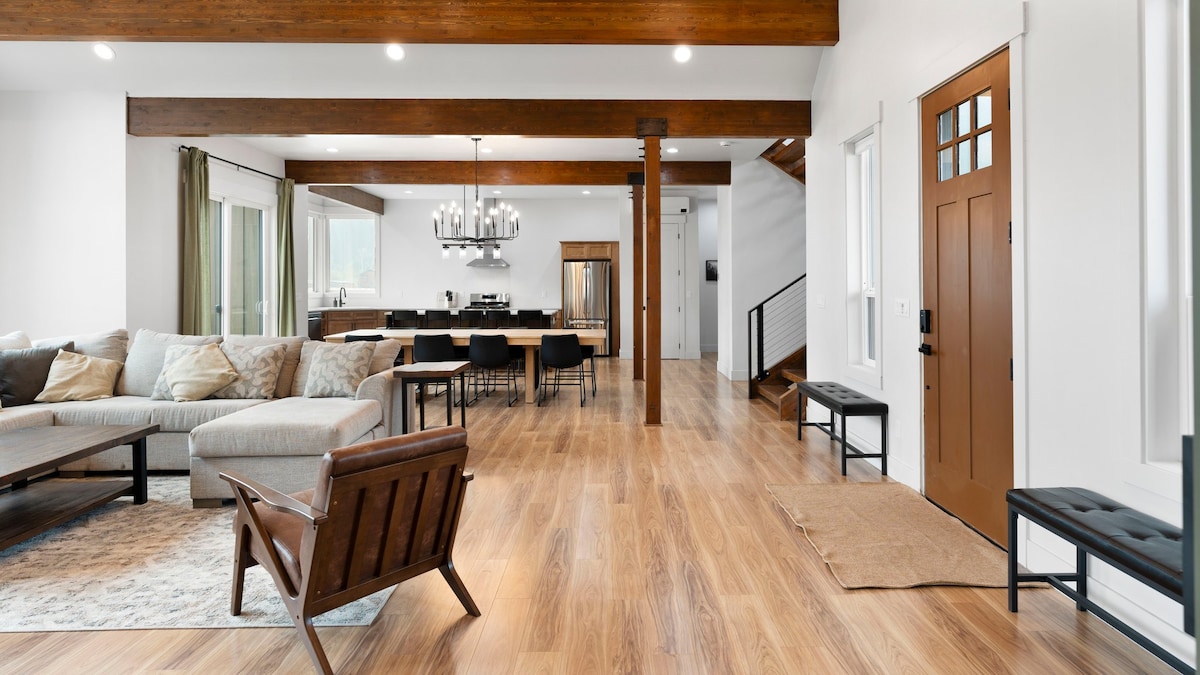 An open-concept living area is highlighted by warm wood beams and abundant natural light. A comfortable sofa and armchairs are arranged around a coffee table. The dining area, featuring a large table with black chairs, flows towards a modern kitchen with sleek appliances.